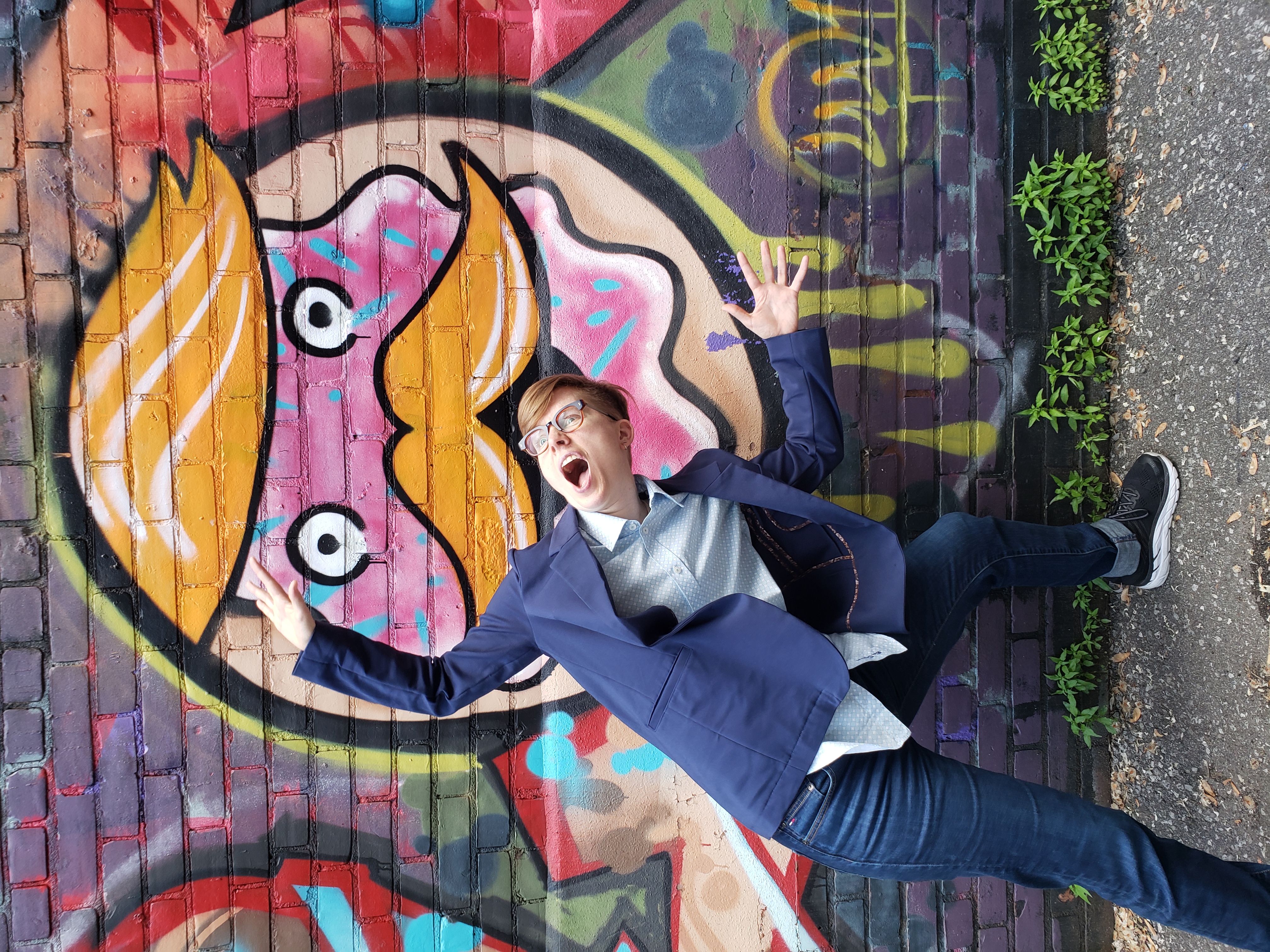 This is an image of Ezra being playful in front of a piece of graffiti. The graffiti looks like a giant donut with a face and Ezra is making a gesture to indicate they might be going to be eaten by the donut.